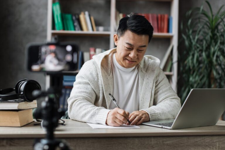 Smiling asian man talking and gesturing in front of modern smartphone camera, writing report while sitting on background of modern home. Live streaming concept.